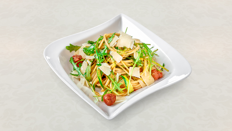 Ein weißer Teller mit Spaghetti, Rucola, Kirschtomaten, Zucchinistreifen und gehobeltem Parmesan auf hellem Hintergrund.