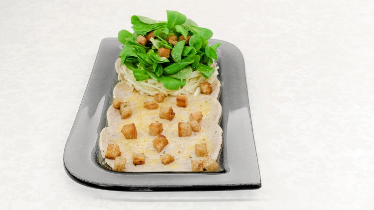 Scheiben von Geflügelfleisch mit Croutons, darauf Feldsalat und Krautsalat in einer schwarzen rechteckigen Schale serviert.