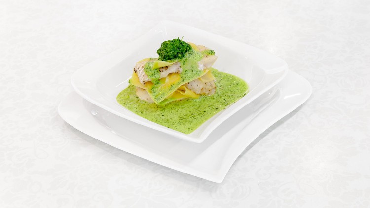 Gestapelte Gemüsestreifen und Fischfilets in grüner Sauce auf einem weißen Teller, dekoriert mit Kräutern.