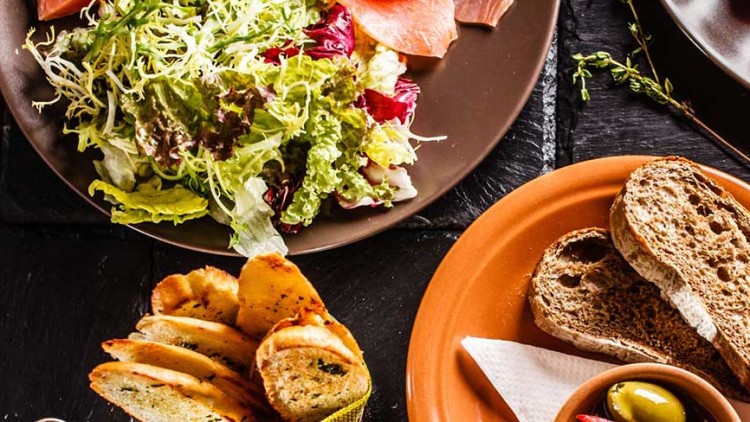 Verschiedene Teller mit Salat, Schinken, Brotscheiben, Oliven, Kräutern und Tomaten-Dip auf dunkler Unterlage.