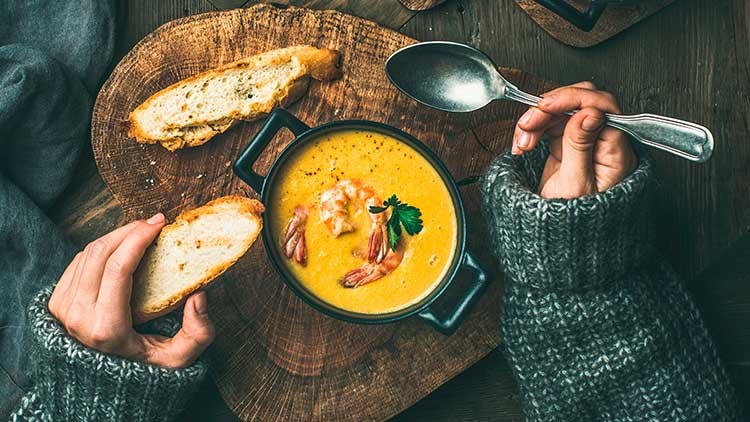 Eine Person hält Brot und einen Löffel, vor ihr steht eine Schale mit gelber Suppe, Garnelen und Petersilie auf einem Holzbrett.