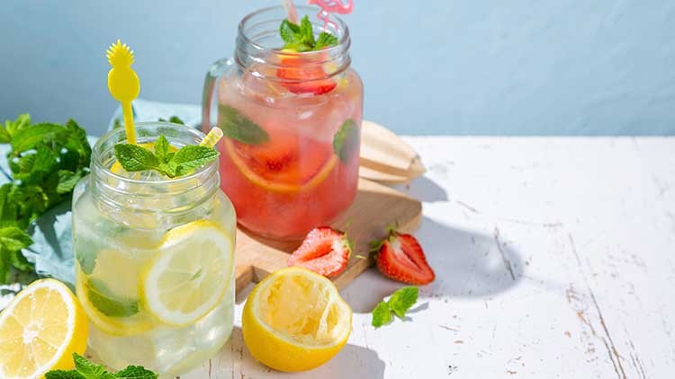 Zwei Gläser mit Zitronen- und Erdbeerlimonade, garniert mit Minze, stehen auf einem hellen Tisch neben Zitronen und Erdbeeren.