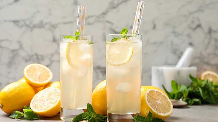 Zwei Gläser Zitronenlimonade mit Eiswürfeln, Zitronenscheiben und Minze, umgeben von Zitronen und Minzblättern, stehen auf grauem Untergrund.