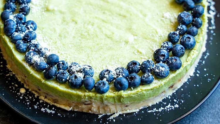 Runder Kuchen mit grünem Belag, garniert mit einer Randdekoration aus frischen Blaubeeren und leicht mit Puderzucker bestreut.