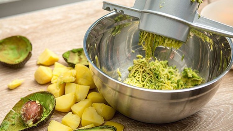 Avocado und Kartoffelstücke liegen neben einer Schüssel auf einem Holztisch, oben eine Presse mit geriebener Avocado.