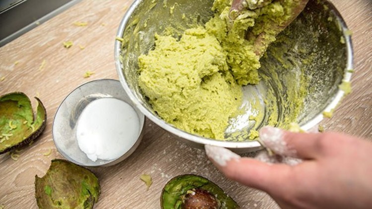 Eine Hand hält eine Schale mit Avocadopüree, daneben liegen Avocadoschalen, Avocadokerne und eine Schale mit weißem Pulver.
