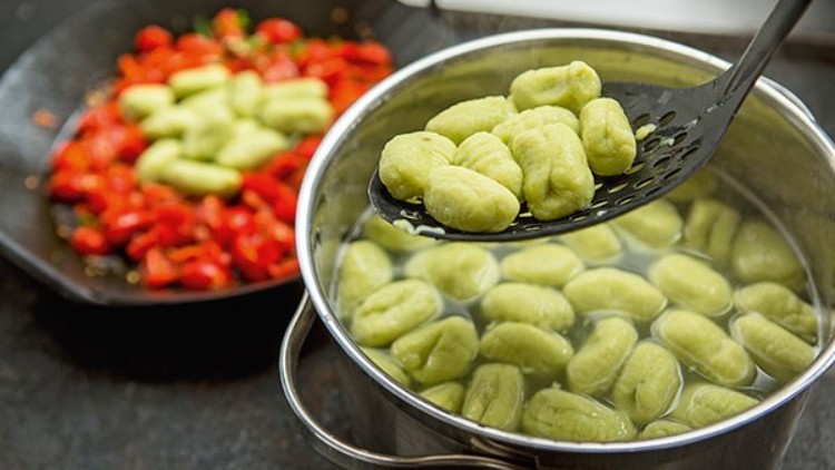 Grüne Gnocchi werden mit einer Kelle aus heißem Wasser gehoben, im Hintergrund eine Pfanne mit Tomaten und weiteren Gnocchi.