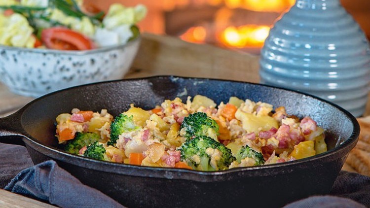 Holztablett mit schwarzer Pfanne voller Gemüse und Speck, daneben ein Salat in einer Schüssel, Toast und Vase, im Hintergrund Kaminfeuer.