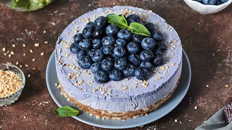 Lila Kuchen mit Blaubeeren, gehackten Nüssen und Minzblättern auf einem Teller, daneben eine Schale mit gehackten Nüssen.
