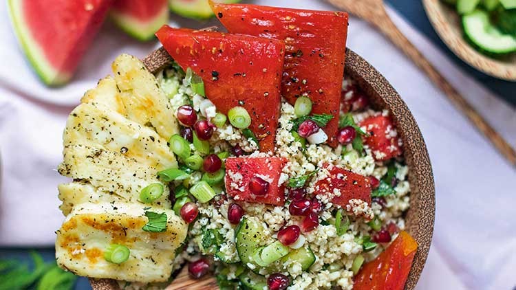 Holzteller mit gegrilltem Käse, Wassermelone, Couscous, Frühlingszwiebeln, Granatapfelkernen und frischen Kräutern.