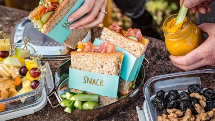 Vollkornsandwiches mit „Snack“-Etikett, Obstspieße, getrocknete Früchte und Dip in Glas mit Gurkenstreifen auf einem Tisch.