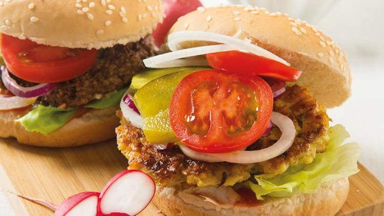 Zwei Burger mit Gemüse, Salat und Sesambrötchen auf Holzbrett, Radieschen und Pommes frites in weißer Schale im Hintergrund.