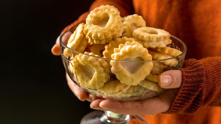Glas mit kurzen Stiel enthält ringförmige, helle Kekse und wird von zwei Händen gehalten, eine trägt orangefarbene Kleidung.