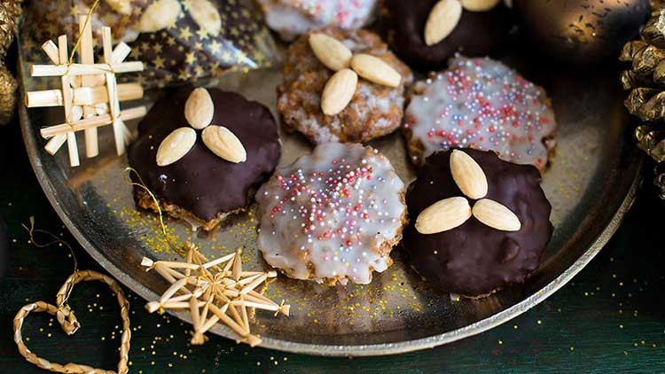 Runde Lebkuchen mit Schokoladen- und Zuckerglasur, verziert mit Mandeln und bunten Streuseln auf einem Metalltablett.