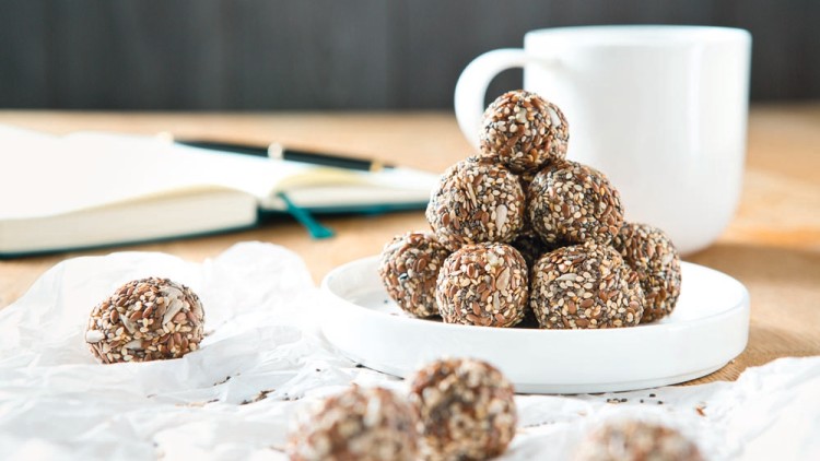 Mehrere Kugeln mit Samen und Körnern liegen auf einem weißen Teller, daneben ein Notizbuch mit Stift und eine weiße Tasse.