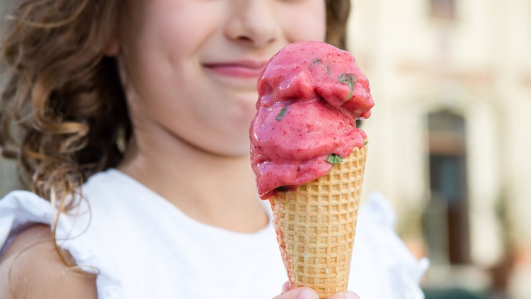 Eine Hand hält eine Eistüte mit zwei Kugeln rotem Eis und kleinen grünen Blättern, im Hintergrund ein unscharfes Gebäude.