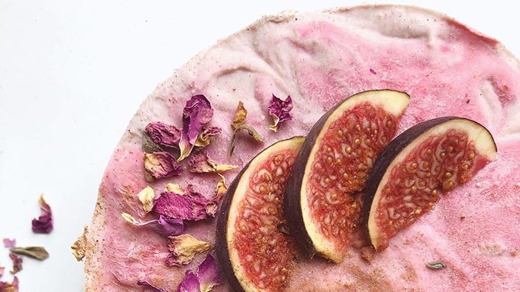 Eine pink-weiße Torte mit getrockneten Rosenblättern und drei Scheiben frischer Feige auf einer hellen Unterlage.