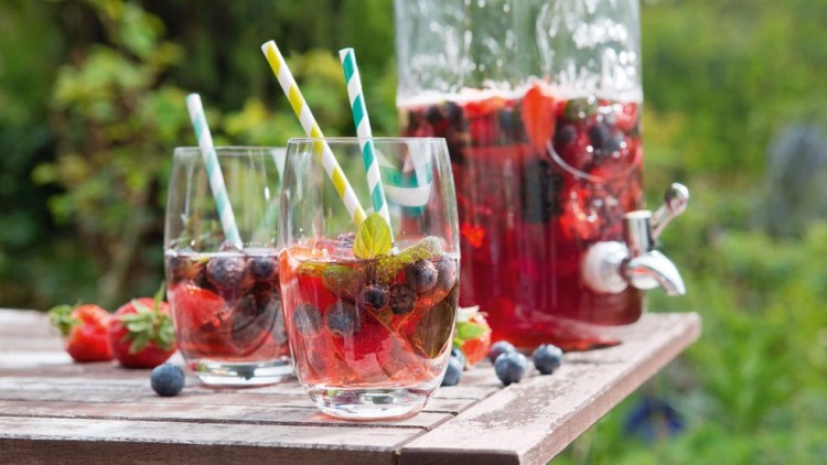 Zwei Gläser mit roten Getränken, Beeren, Minze und gestreiften Trinkhalmen stehen auf einem Holztisch, daneben ein Getränkespender.