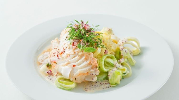 Ein weißer Teller mit gedämpftem Fischfilet, gekochtem Lauch, Senfsauce, Kartoffeln und frischen Kräutern als Garnitur.