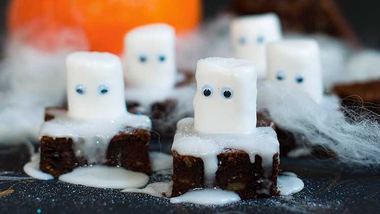 Weiße Marshmallows mit Augen stehen auf Schokobrownies, umgeben von weißer Glasur und Dekoration in Spinnennetzoptik.