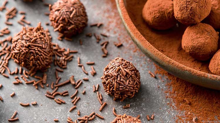 Auf einem grauen Untergrund liegen Schokotrüffel mit Schokostreuseln, daneben eine Schale mit Trüffeln in Kakaopulver.