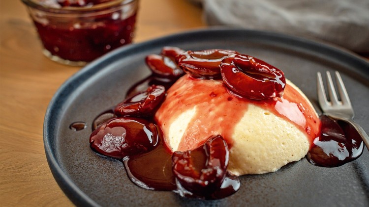 Grießpudding auf einem dunklen Teller, garniert mit roten heißen Pflaumen und Soße, daneben eine kleine Gabel und ein Glas Pflaumenkompott.