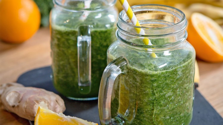 Zwei Gläser mit grünem Smoothie und gelb-weiß gestreiften Strohhalmen, daneben Ingwer und ein Stück Orange.
