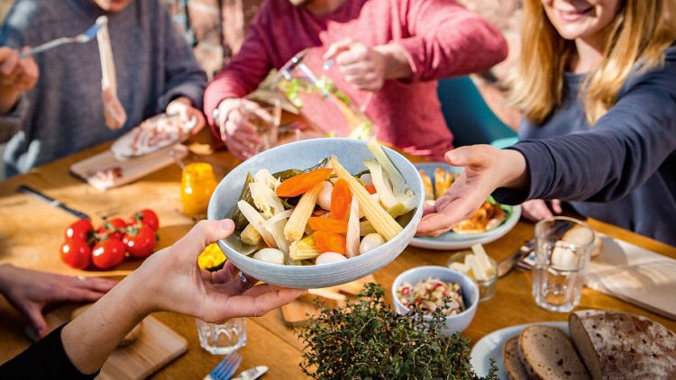 Person reicht eine Schüssel mit verschiedenen eingelegten Gemüsesorten an einem gedeckten Tisch mit Brot, Salat und Getränken weiter.