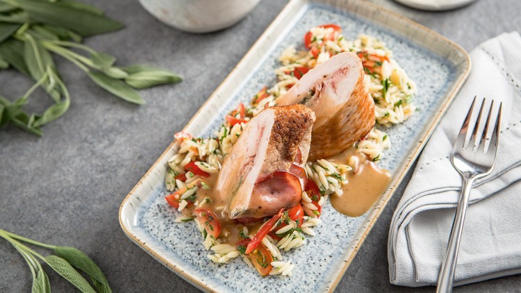 Gefüllte Hähnchenbrust mit Sauce, Orzo-Nudelgemüse und frischen Kräutern auf einem länglichen Teller neben Besteck und Serviette.