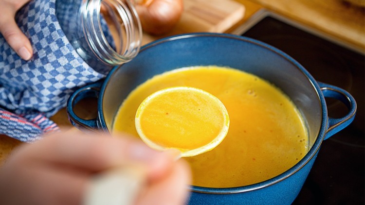 Eine Person hält ein Einmachglas mit Tuch und schöpft gelbe Suppe mit einer Kelle aus einem blauen Kochtopf.