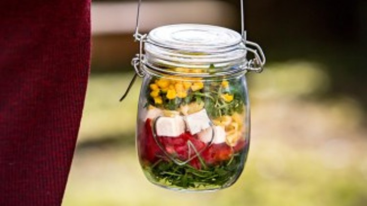 In einer Glasvorratsdose sind frische Salatzutaten wie Rucola, Tomaten, Käsewürfel und gelbe Blüten zu sehen.