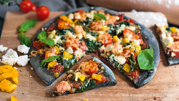 Pizza mit schwarzem Boden, gelber Paprika, Tomaten, Blattspinat, Käse und Basilikum auf Holzbrett mit Dekoration.