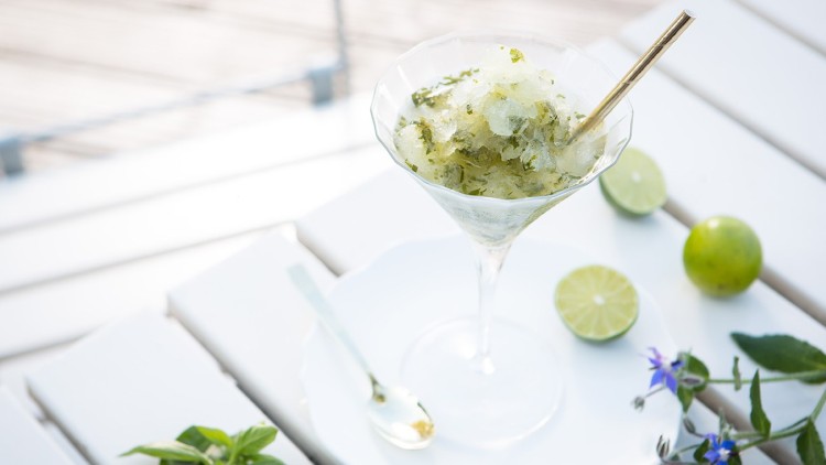 Ein Glas mit grünem Sorbet und goldfarbenem Strohhalm steht auf einem weißen Tisch mit Limettenhälften und Kräutern.