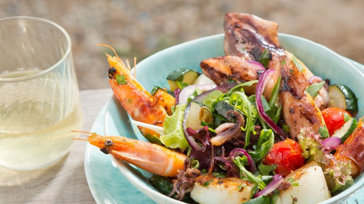 Schale mit Grilltintenfisch, Garnelen, Zucchini, Salat, Tomaten und Zwiebeln neben einem Glas Weißwein auf Holztisch.