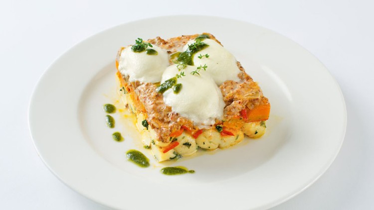 Ein Stück Gemüse-Lasagne mit geschmolzenem Mozzarella und grüner Kräutersoße auf einem weißen Teller.