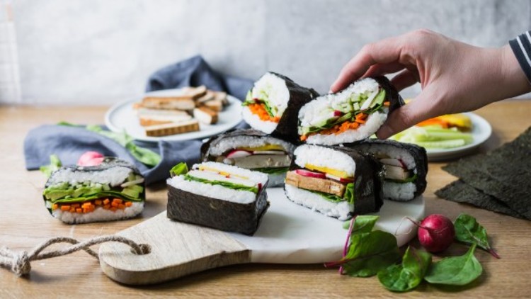 Hand nimmt ein halbiertes Onigirazu mit Nori, Reis, Gemüse und Tofu von einem Holzbrett, daneben Radieschen und Spinat.