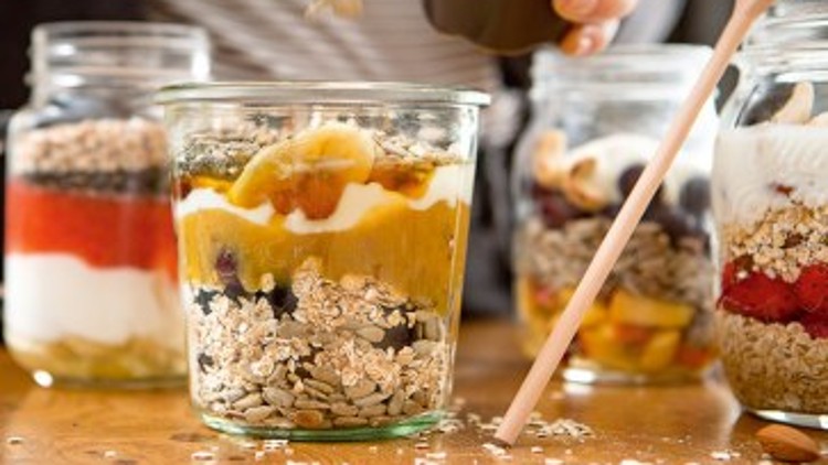 Eine Hand streut Körner in ein Glas mit Joghurt, Früchten und Müsli, weitere befüllte Gläser stehen auf einem Holztisch.