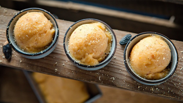 Drei Schalen mit orangefarbenem Sorbet, bestreut mit geriebener dunkler Schokolade, stehen auf einer Holzoberfläche.