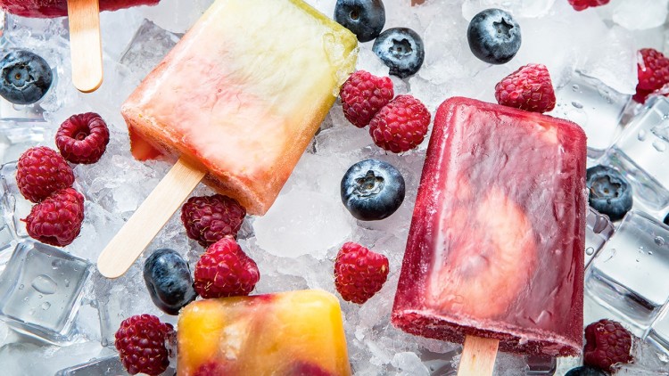 Gefrorenes Fruchteis am Stiel in verschiedenen Farben liegt auf Eiswürfeln, umgeben von Himbeeren und Heidelbeeren.