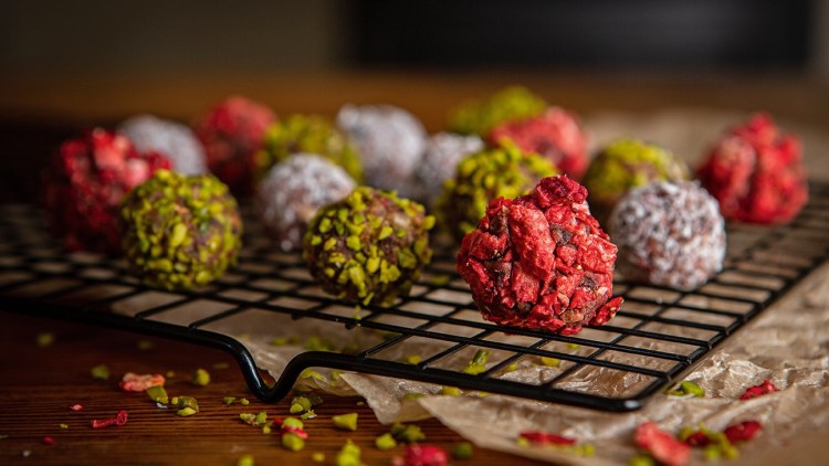 Bunte Pralinen auf einem schwarzen Gitter, dekoriert mit roten Beerenstücken, grünem Pistaziengranulat und Puderzucker.
