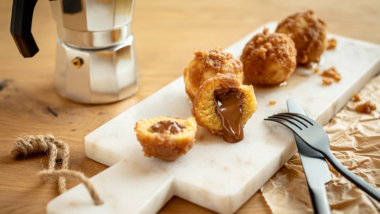 Frittierte Gebäckstücke mit flüssiger Schokoladenfüllung auf weißem Brett, daneben Besteck und silberne Espressokanne.