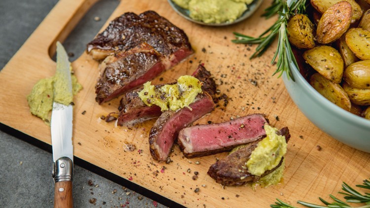 Medium-rare gebratenes Steak in Scheiben, serviert mit Kräuterbutter, Rosmarin, Ofenkartoffeln und einer kleinen Schale Dip.