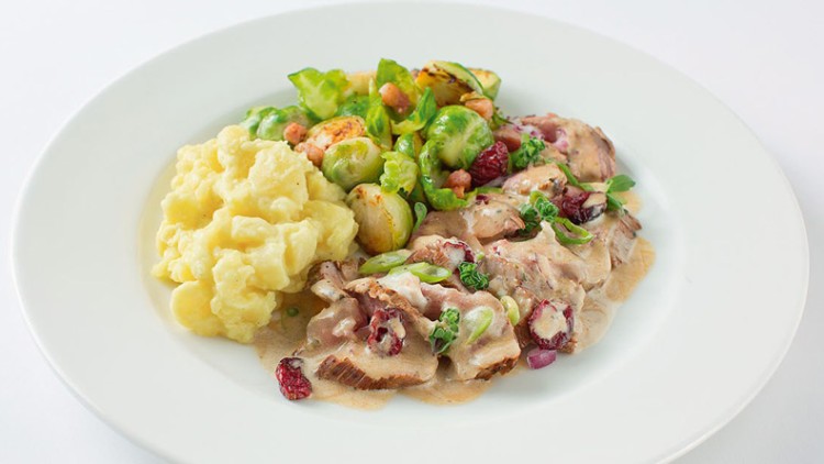 Gericht auf weißem Teller mit Kartoffelpüree, Rosenkohl und Fleischscheiben in heller Soße mit Kräutern und Preiselbeeren.