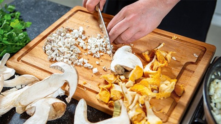 Eine Hand schneidet Pilze auf einem Holzbrett, daneben liegen weitere Pilze, Kräuter und eine Pfanne mit Zwiebeln.