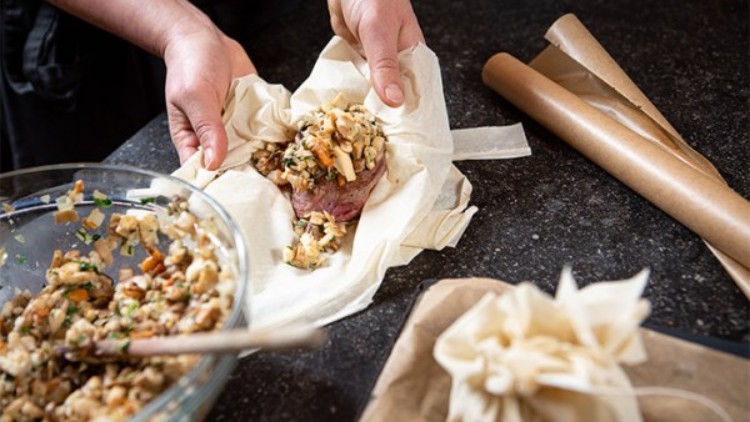 Hände legen eine Füllung aus Kräutern, Gemüse und Brot auf ein Stück Fleisch, das in Backpapier auf einer Arbeitsfläche liegt.