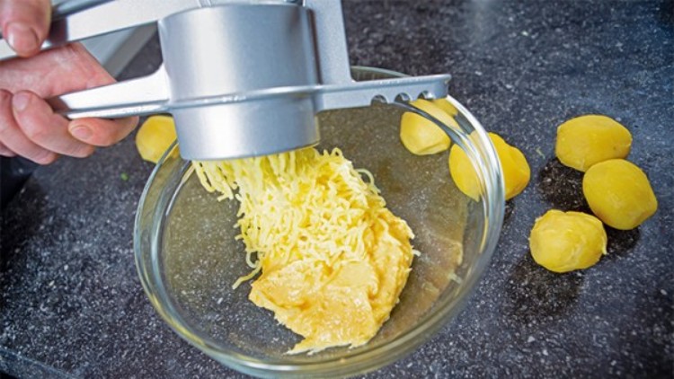 Gekochte Kartoffeln werden mit einer silbernen Kartoffelpresse in eine Glasschüssel gepresst, weitere Kartoffeln liegen daneben.