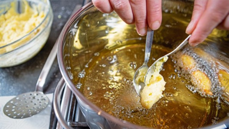 Eine Person formt mit zwei Löffeln Teigbällchen, die in heißem Fett in einem Topf auf einem Herd frittiert werden.