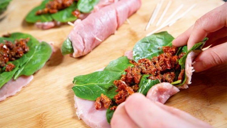Hand rollt eine Scheibe Schinken mit frischem Blattspinat und getrockneten Tomaten auf einem Holzbrett.