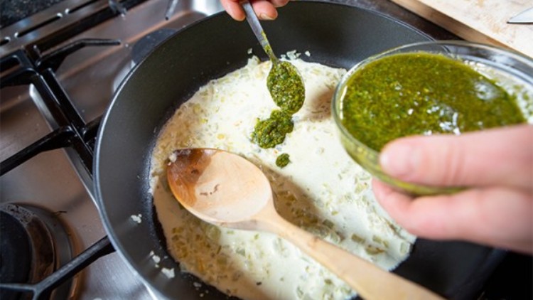 Eine Pfanne mit heller Sauce auf einem Herd, in die Pesto aus einer Schale mit einem Löffel hinzugefügt wird.