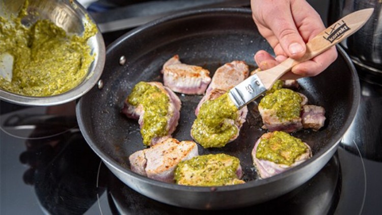 Runde Fleischstücke in einer Pfanne werden mit einem Pinsel und grünem Pesto bestrichen.